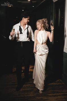 a man and woman standing next to each other holding wine glasses in their hands as they walk down the hall