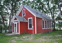 a small red house with an american flag on the roof and two windows, in front of some trees