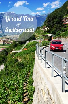 a red car driving down a road next to a lush green hillside with mountains in the background