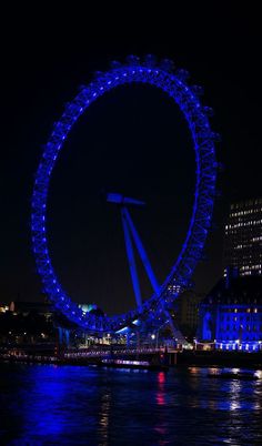 the london eye lit up in blue at night with lights reflecting off water and buildings behind it