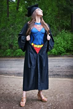 a woman in a graduation gown and cap is looking up at the sky while wearing a superman cape