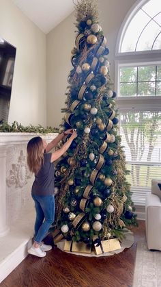 a woman decorating a christmas tree with gold and silver ornaments on the top, in front of a large window