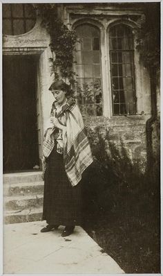 an old black and white photo of a woman standing in front of a house wearing a shawl