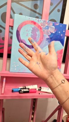 a person's hand is holding up a painting in front of a pink shelf