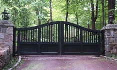 an iron gate in front of a stone wall and tree lined driveway with two lamps on each side