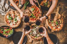 people are eating pizza and drinking wine at a table full of different types of food