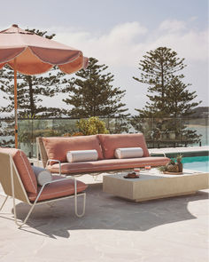 Elegant outdoor seating area showcasing a 3-seat sofa and armchairs in a soft pink hue with a matching umbrella. The setup is styled with tropical fruits and drinks, capturing a relaxed, upscale vibe near the coast. From Business & Pleasure Co. x Coco Republic collaboration. Aesthetic Signature, 3 Seat Sofa