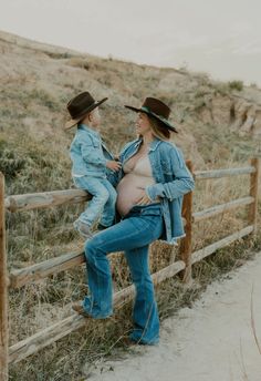 a pregnant woman sitting on top of a wooden fence next to a man wearing a cowboy hat