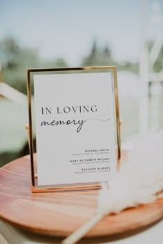 a sign that says in loving memory sitting on top of a wooden table next to a white feather