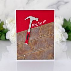 a red and white card with a hammer stuck in it on a table next to flowers