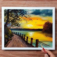 a person is holding a paintbrush while painting a sunset on a wooden deck by the water