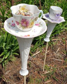 two white cups and saucers sitting on top of a stand in the grass next to flowers