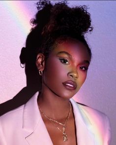 a woman wearing a white suit and gold necklace standing in front of a rainbow colored wall