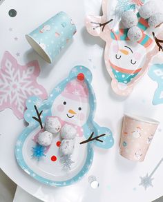 a white table topped with plates and cups filled with snowman themed food next to cupcakes