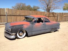 an old car is parked in the dirt near a wooden fence and some trees with no leaves on it