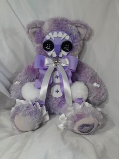 a purple teddy bear with big eyes sitting on a white bed sheet and wearing a purple ribbon around its neck