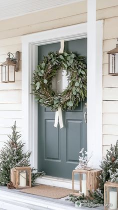 a wreath on the front door of a house with two small christmas trees and lanterns