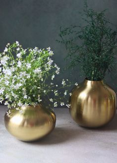 two gold vases with white flowers in them on a gray surface next to each other