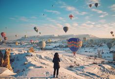 a woman standing in the snow looking at hot air balloons flying over her and mountains