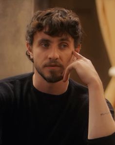 a man sitting down with his hand on his chin and looking at the camera while wearing a black t - shirt