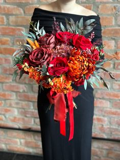 a woman wearing a black dress holding a red and orange bouquet in front of a brick wall