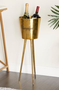 two bottles of champagne are in a gold ice bucket on a stand next to a potted plant