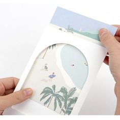 two hands holding an open card that has a beach scene on it and palm trees in the background