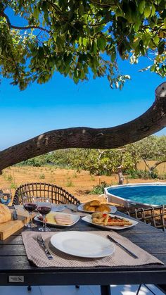 an outdoor dining table with food and wine on it, under a tree overlooking the ocean