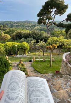 an open book sitting on top of a grass covered field next to a tree filled hillside