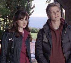 a man and woman standing next to each other in front of some trees with the ocean in the background