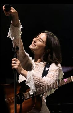 a woman holding a guitar and singing into a microphone with her right hand in the air