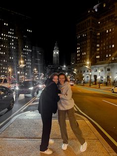 two women standing on the sidewalk in front of some buildings at night with their arms around each other