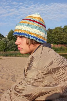 a young man sitting on the beach wearing a multicolored knitted beanie