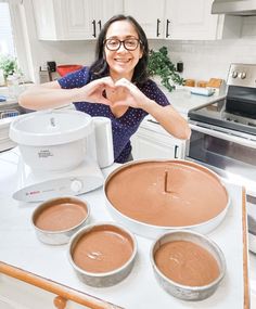 a woman standing in front of three pans of cake batter and making heart shape with her hands