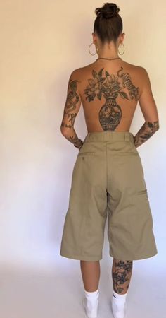 a woman with tattoos standing in front of a white wall wearing khaki shorts