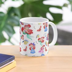 the little mermaid mug is sitting on top of a wooden table next to a book