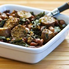 a white bowl filled with meat and beans on top of a wooden table next to a knife