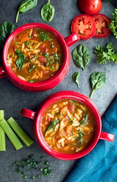 two red bowls filled with vegetable soup on top of a blue cloth next to tomatoes, celery and green beans