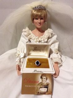 a barbie doll wearing a tiara and holding a ring in a box on a bed