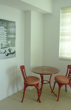 two red chairs and a round table in a room with white walls, beige flooring and windows