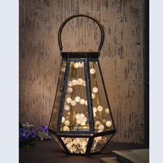 an old fashioned lantern with lights in it on a table next to flowers and a book