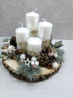 some white candles are sitting on a piece of wood with pine cones and greenery