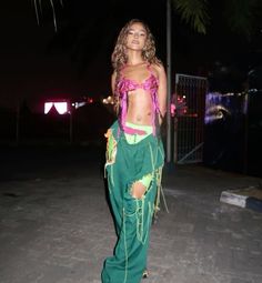 a woman in a pink top and green pants is standing on the street at night