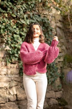 a woman standing in front of a stone wall wearing a pink sweater and white pants