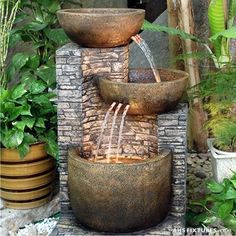 a water fountain with three bowls on top and two jets running down the side, surrounded by greenery