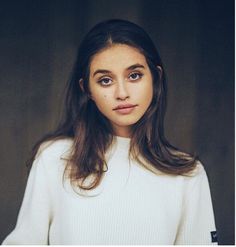 a woman with long brown hair wearing a white sweater and looking at the camera while standing in front of a dark background