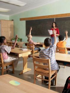 several children are sitting at desks and raising their hands in front of the teacher