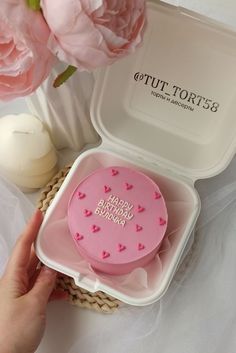 a person holding a pink cake in a plastic container with flowers and candles behind it