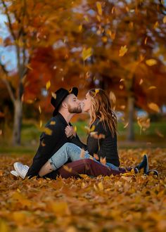 a man and woman sitting on the ground kissing in front of trees with falling leaves