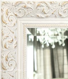 an ornate white framed mirror with gold trimmings on the edges and a chandelier in the background